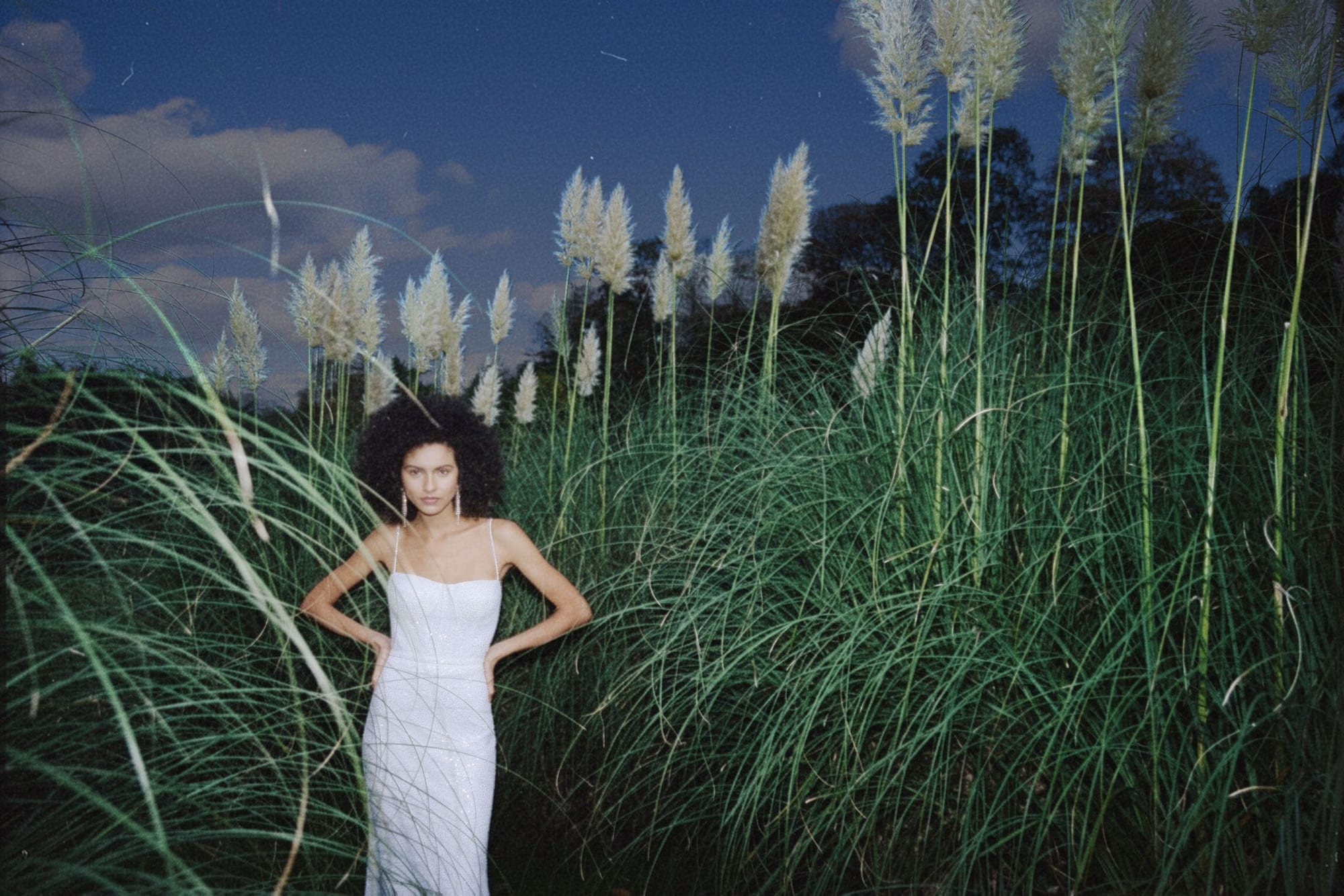 A bride in a white slip dress stands amid tall pampas grass at dusk, hands on her hips, meeting the camera's gaze