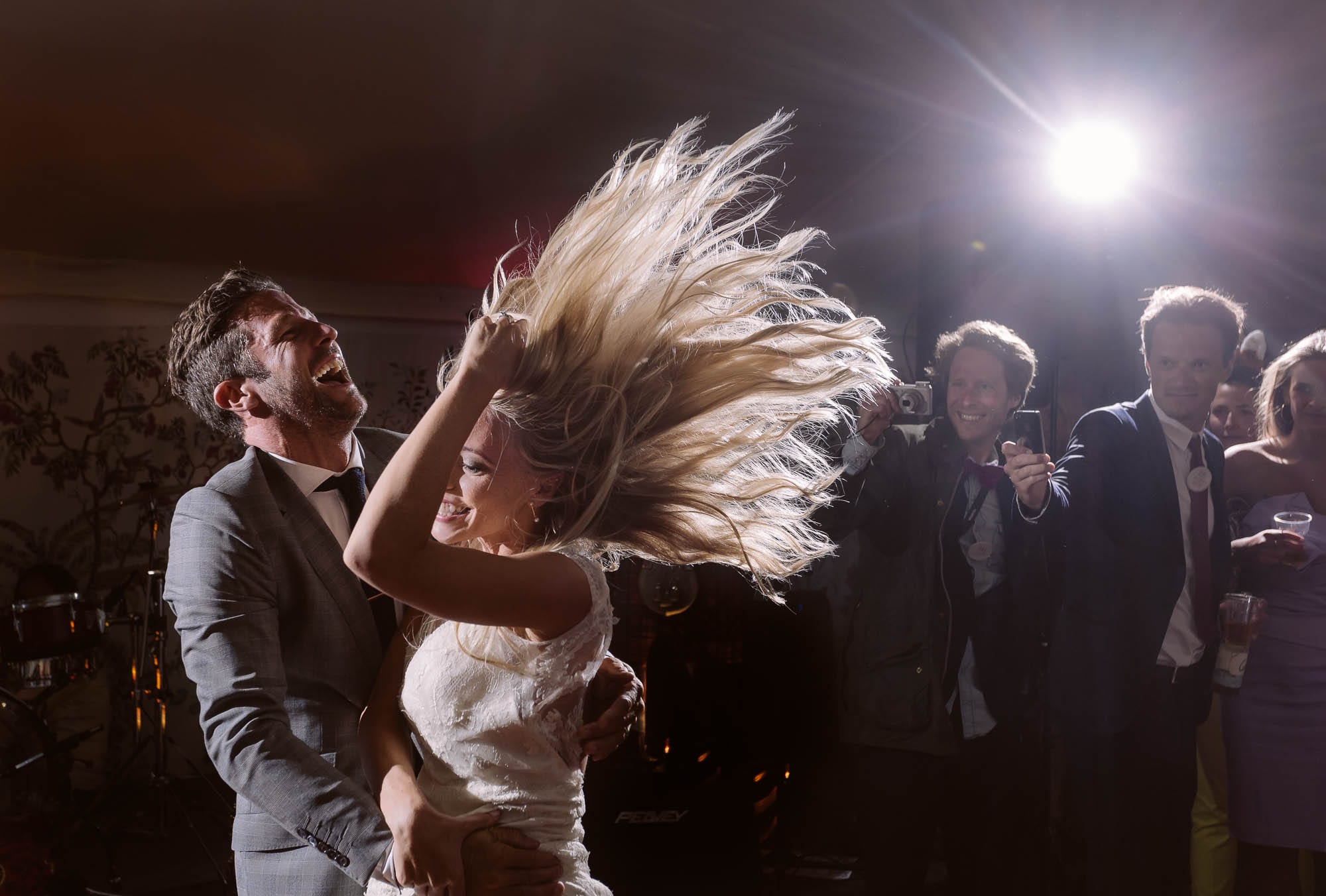 A bride's hair flies mid-dance, backlit by flash, while laughing guests photograph the moment